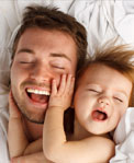 Close view of laughing father and toddler lying in bed with baby's hands on dad's face